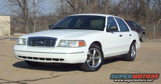 White sedan with aftermarket wheels parked on asphalt, front quarter view. Another vehicle is visible behind a fence