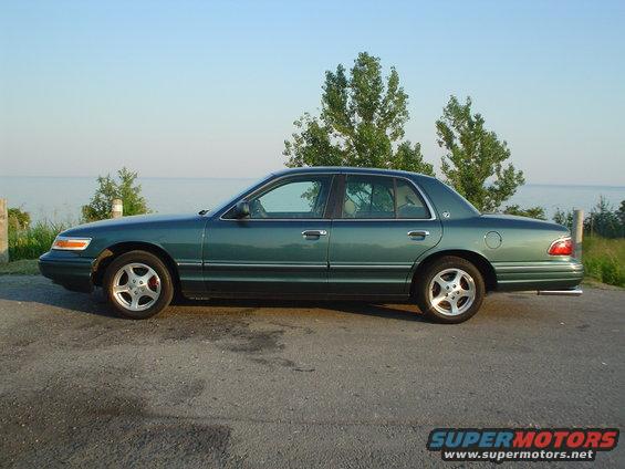 Green sedan parked on asphalt with water and trees in the background