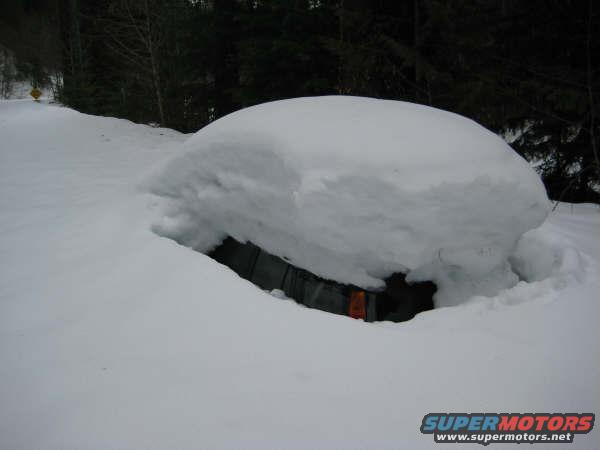 Car covered in deep snow with only a bit of the front fender and headlight visible