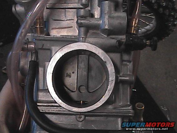 Motorcycle carburetor with a circular intake and visible throttle plate. A chain and spokes are visible in the background