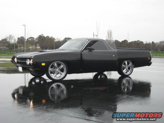 Black 1970 Ford Ranchero parked on wet asphalt, reflecting in puddles