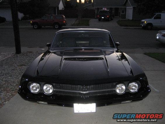 Front view of a black coupe with a hood scoop parked in a driveway. Other vehicles and houses are in the background