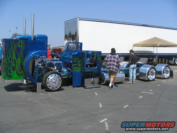 Blue custom semi-truck chassis with green flames, chrome fenders, and two people standing nearby