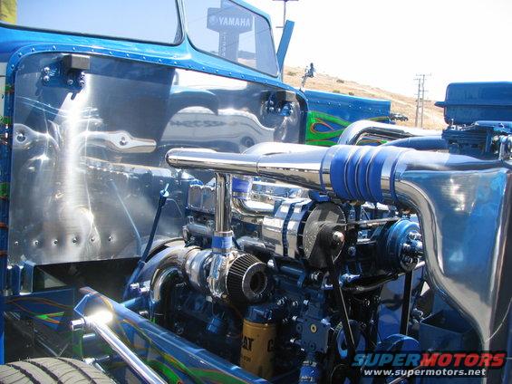 Close-up of a blue customized truck engine with chrome exhaust pipes and a Yamaha sign reflected in the door