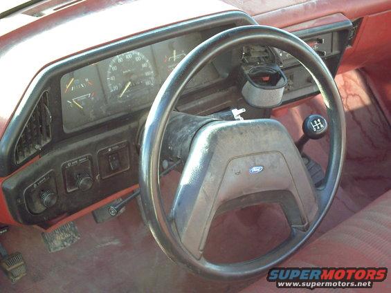 Truck interior with a worn steering wheel, dashboard, and manual gear shifter. Red upholstery is visible