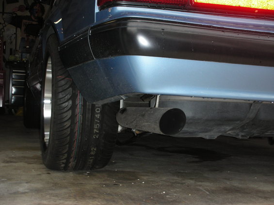 Blue car's rear bumper and exhaust pipe, showing a black bumper trim and a large, dark exhaust pipe. Tire sidewall text is visible