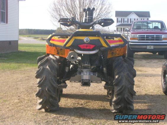 Rear view of a yellow and red quad bike with large mud tires. A red pickup truck is parked behind it