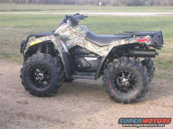 Camouflaged Can-Am Outlander ATV with large mud tires parked on dirt