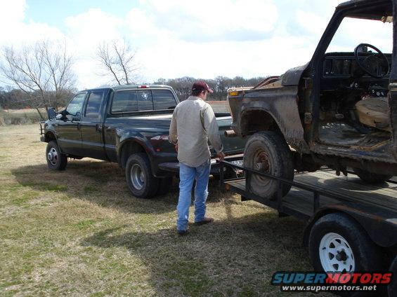 Dark green pickup truck towing a rusty, stripped-down vehicle on a trailer