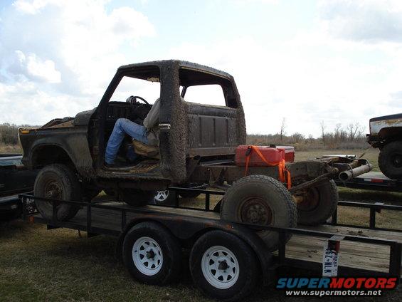 Rusty truck chassis on a trailer, viewed from the side, with a person sitting in the driver's seat