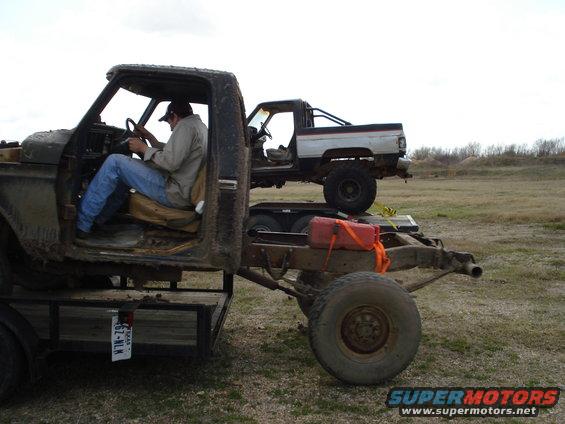 Man sits in cab of old truck on trailer; another truck is on top of it