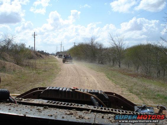 Several vehicles driving down a dusty dirt road with trees and utility poles lining the sides