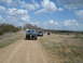 2008 International School Bus - A convoy of vehicles, led by a blue pickup truck, travels down a dusty rural road under a cloudy sky.