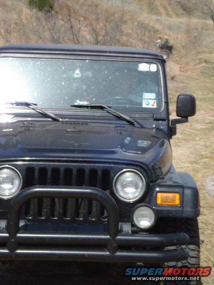 Dark blue Jeep Wrangler front grille with bull bar, dusty windshield, and a distant ATV on a hill