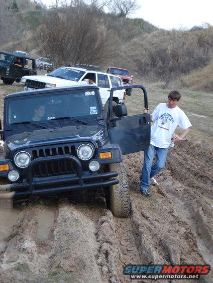 Jeep Wrangler stuck in mud, driver's door open, with a white SUV and red truck behind it