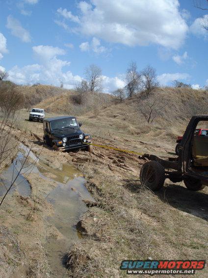 Black Jeep Wrangler stuck in mud, being pulled by a yellow tow strap attached to an unseen vehicle. Another white SUV is behind it