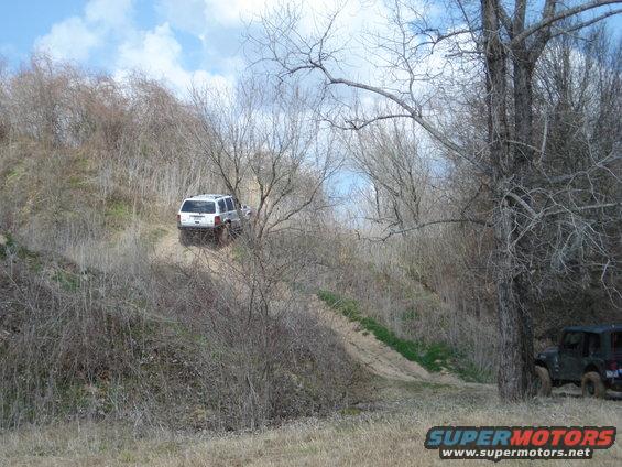 White SUV climbing a dirt hill off-road, green Jeep parked to the right