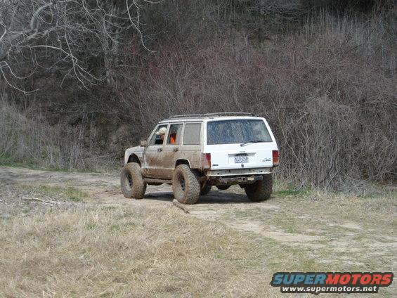 White Jeep Cherokee with big tires driving on a dirt path, covered in mud. Bare trees and brush in background