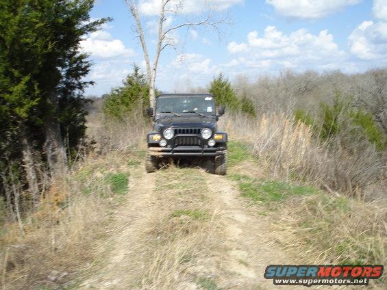 Black Jeep Wrangler on a dirt road, front view. The vehicle has a brush guard and is surrounded by dry grass and trees