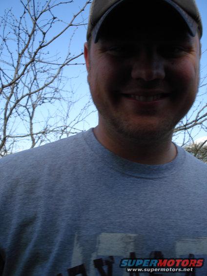 man in a gray t-shirt and baseball cap smiling outdoors with bare tree branches and blue sky behind him. Text on shirt reads SUPERMOTORS