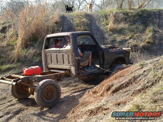 Mud-covered pickup truck chassis with two people inside, stuck on a dirt hill. Red gas can on the rear