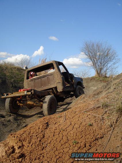 Mud-covered truck chassis with exposed frame and rear axle, driving up a dirt embankment under a blue sky