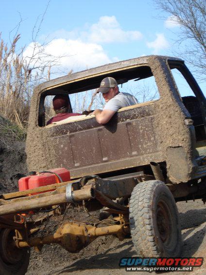 Mud-covered truck chassis with two people in the cab, red gas can mounted on frame