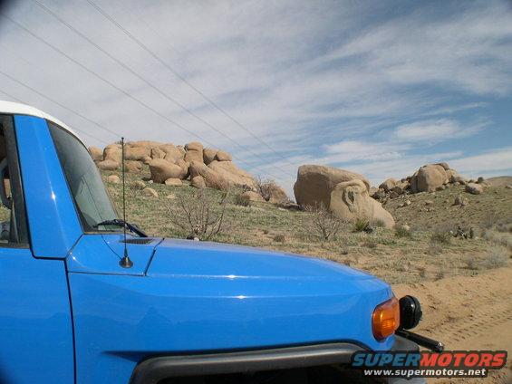 Blue Toyota FJ Cruiser front fender and hood with a white roof, parked on a dirt road with rocky hills in the background