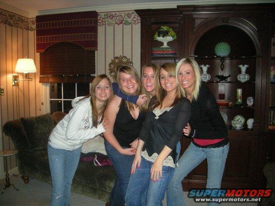 Five young women posing for a photo indoors, with a couch and bookshelf in the background