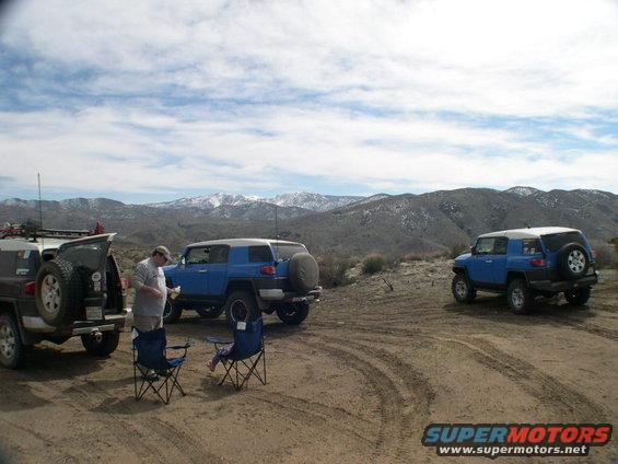 Three blue Toyota FJ Cruisers parked on a dirt track, with snow-capped mountains in the distance. Two camping chairs are set up