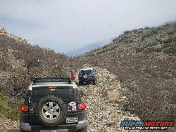 Two Toyota FJ Cruisers driving on a rocky, uphill trail with sparse vegetation