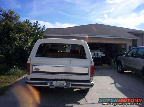 Rear view of a white Ford Bronco parked in a driveway. Another SUV is parked to the right, and a Jeep is visible in the garage