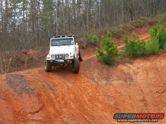 White Jeep Wrangler driving up a red dirt hill with trees in the background