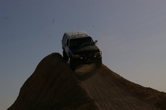 Dark SUV with white camper shell driving up a dirt mound, dust kicking up