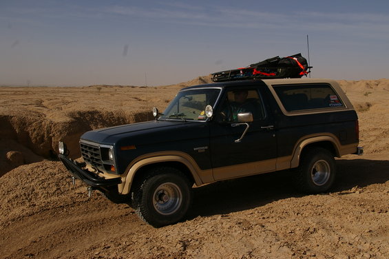 Dark blue and tan Ford Bronco with a roof rack loaded with gear, driving on a dirt track in a desert landscape