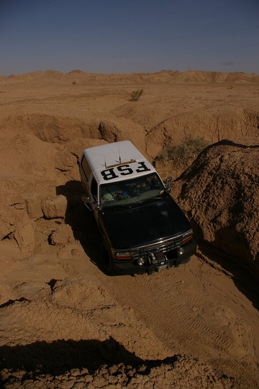 1986 Ford Bronco with a white camper shell navigating a sandy desert wash. Text 'FSB' is visible on the shell