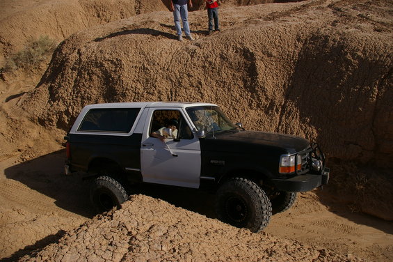 Black and white Ford Bronco driving through dirt hills. Two people stand on a ridge above