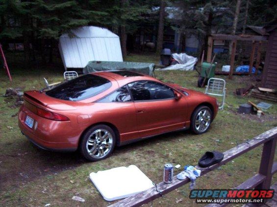 Orange sports coupe parked in a yard with trees and a shed. Sunroof is open