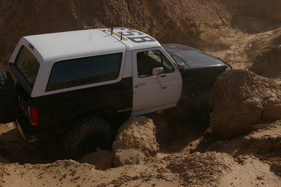 Two-tone Ford Bronco navigating a sandy, rocky off-road trail. Numbers '358' are visible on the roof