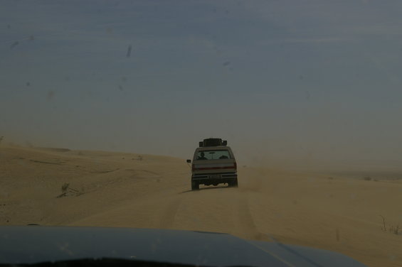 White SUV driving on a dirt road in a desert, with luggage on its roof rack