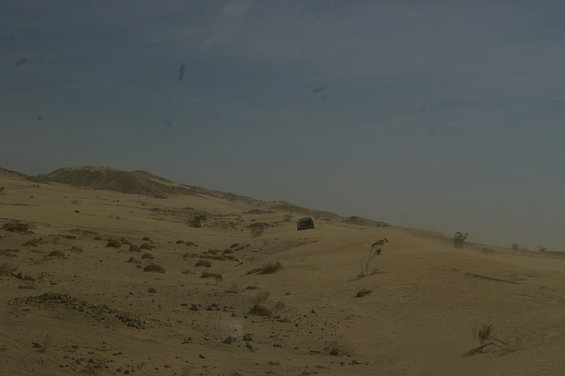 Desert terrain with two dark-colored vehicles driving on a sandy ridge under a hazy blue sky