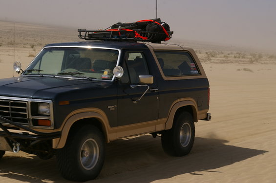 Dark blue and tan Ford Bronco with roof rack loaded with gear, driving on a sandy terrain