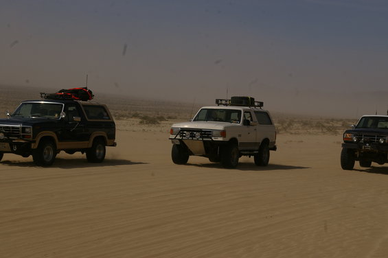 Three Ford Broncos driving in a desert landscape. The lead vehicle is white, the second is dark blue, and the third is dark blue