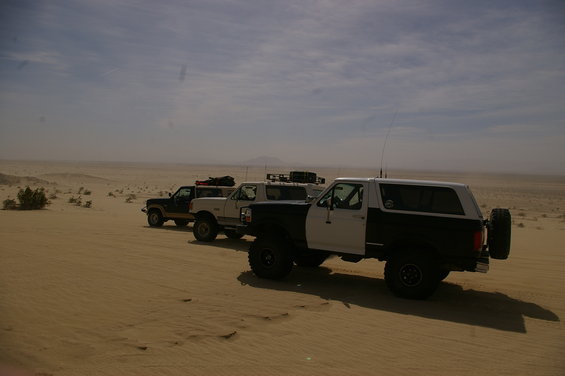 Three trucks and a Bronco parked on a sand dune under a hazy sky