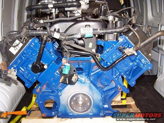 Blue painted V8 engine block with wiring harnesses and hoses attached, sitting on a wooden pallet inside a van