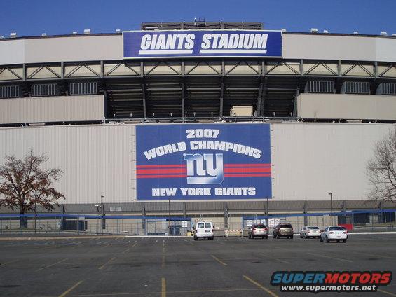 Giants Stadium exterior with 'GIANTS STADIUM' and '2007 WORLD CHAMPIONS NEW YORK GIANTS' banners; several cars and a van parked in lot