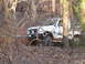 1987 Suzuki Samurai - A white 1987 Suzuki Samurai navigates a wooded trail, its tires gripping the uneven terrain.