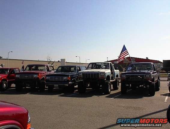 Several Ford Broncos and F-Series trucks parked in a lot, with an American flag flying in the background