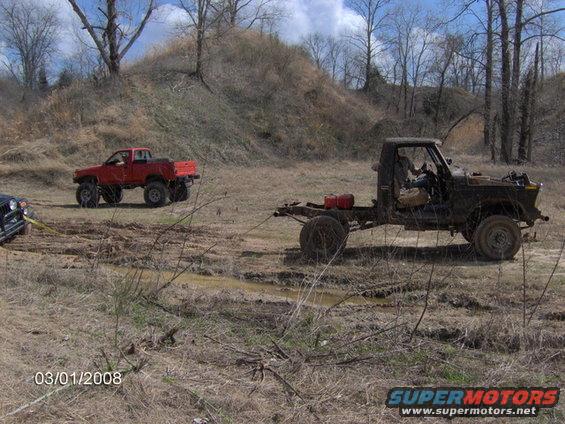 Dark, boxy, open-top vehicle with a driver, parked on dirt near a red car