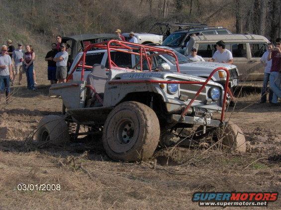 Modified off-road buggy with roll cage stuck in mud, other vehicles and people in background